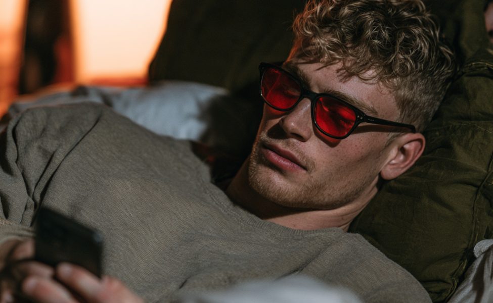 Mann liegt abends im Bett mit dem Smartphone und trägt eine Brille mit roten Gläsern