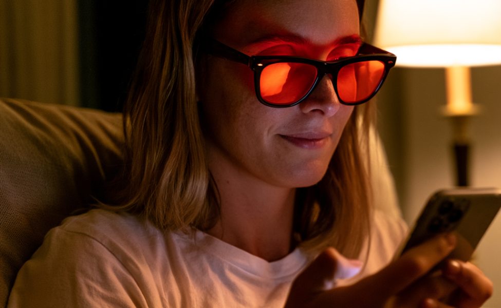 Frau schaut abends auf ihr Smartphone neben einer warm leuchtenden Nachttischlampe